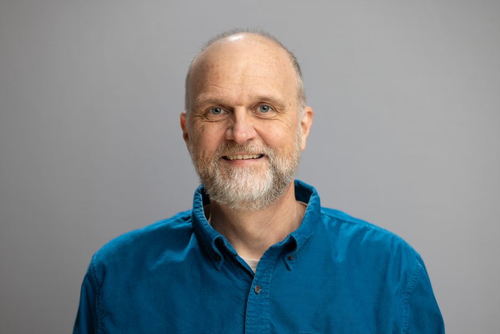 Tom smiles. Tom wears a royal blue button up.