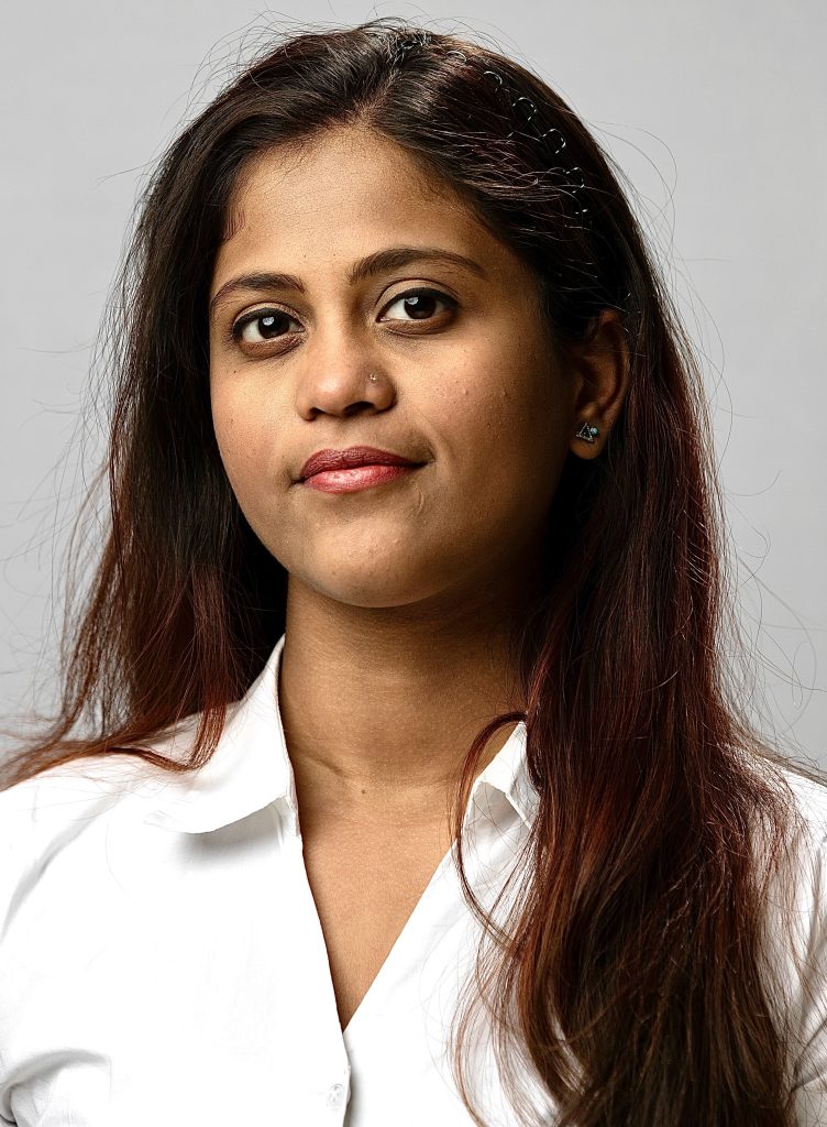 Gopika smiles. She has long, dark brown hair and wears a white button up.
