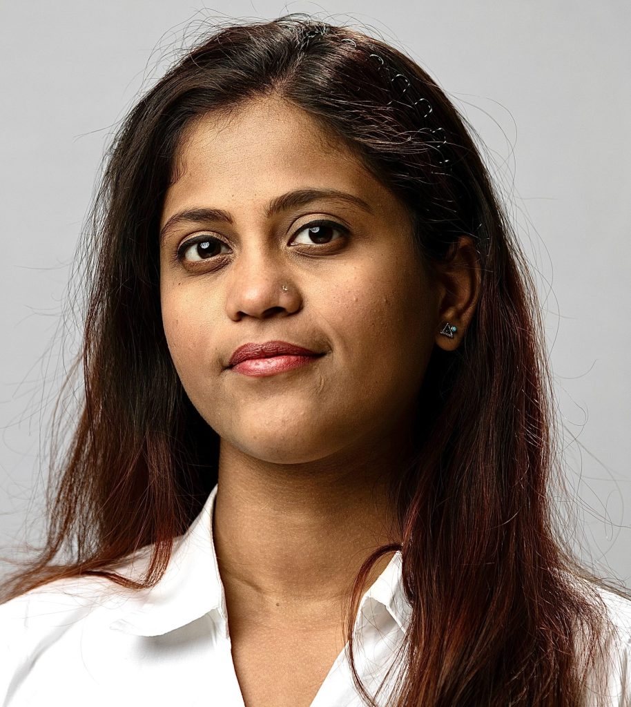 Gopika smiles. She has long, dark brown hair and wears a white button up.