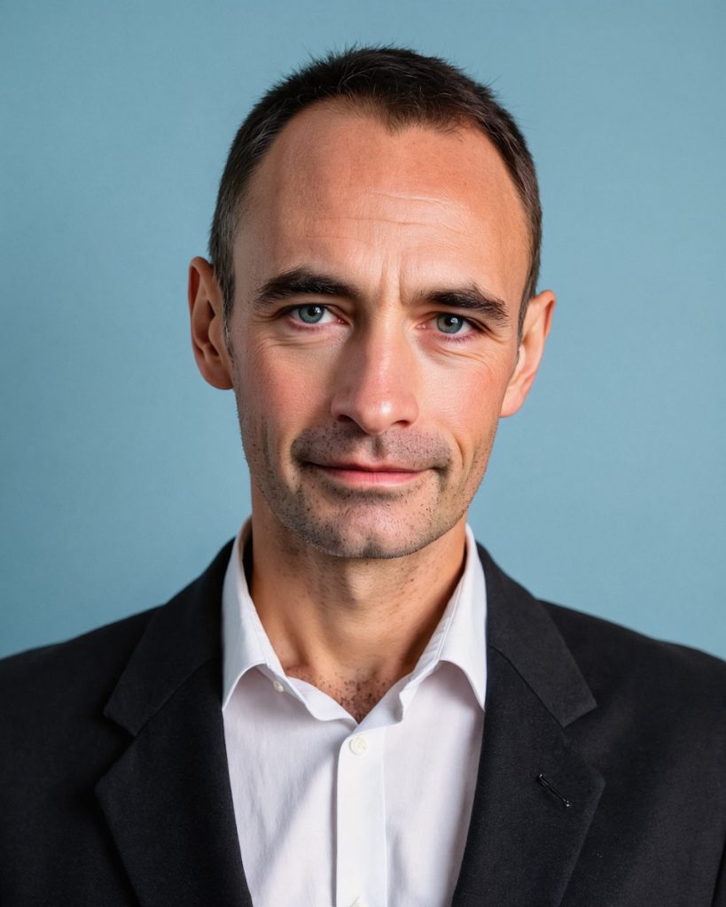 Jean-Michel smiles. He has short, dark hair and wears a white button up and suit.