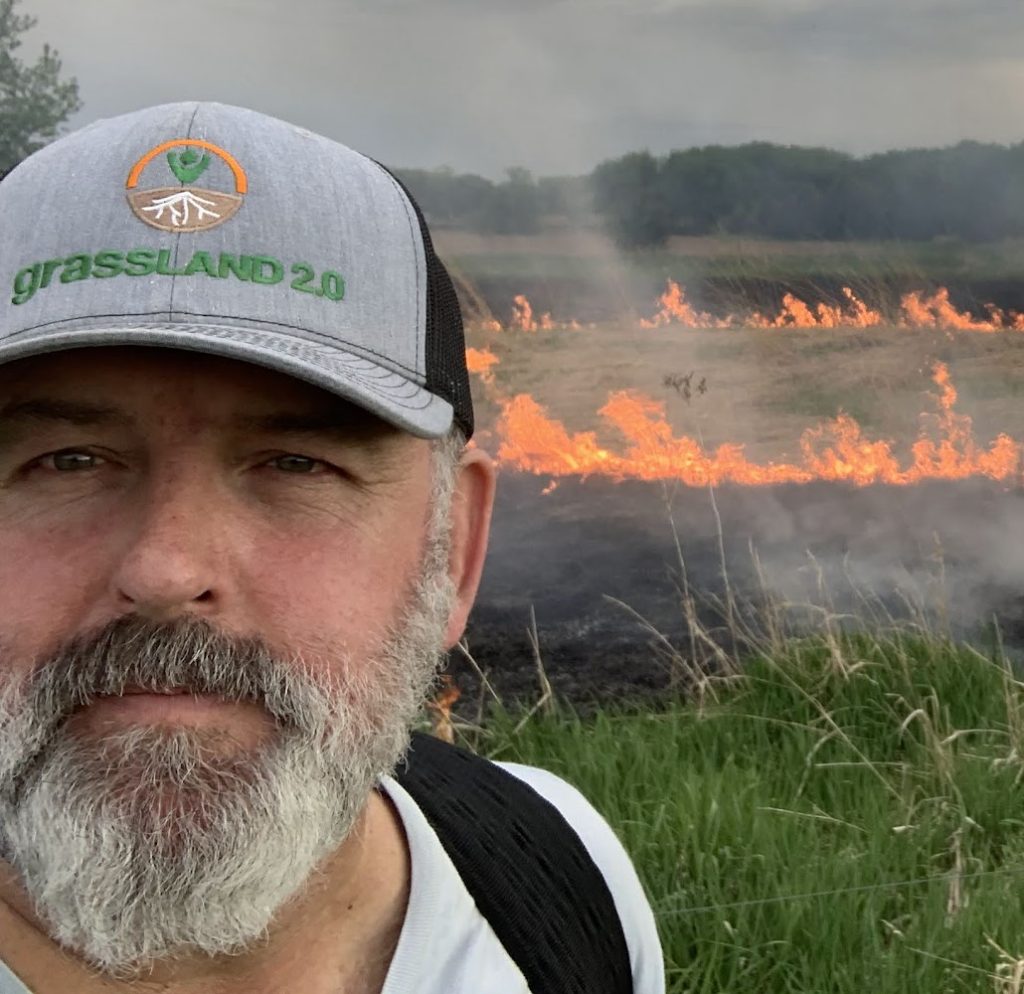 Randy smiles ever so slightly. Randy wears a baseball cap that says "grassland 2.0" and stands in front of a prairie fire.
