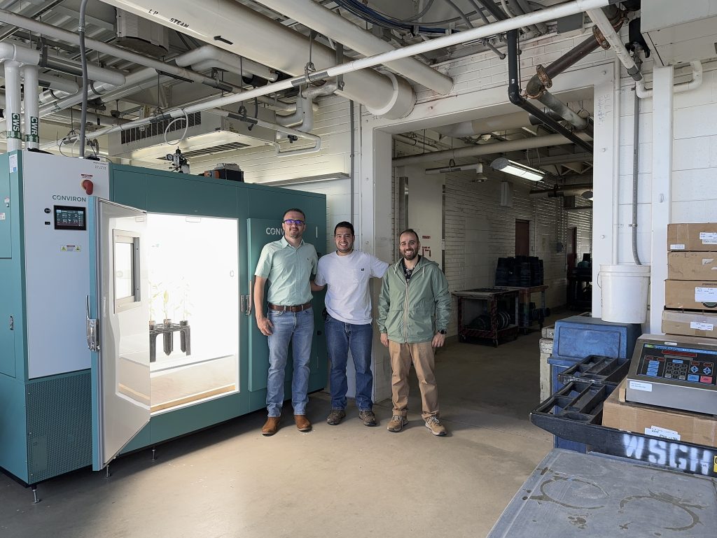 Three people pose for a picture and smile in front of a large growth chamber.