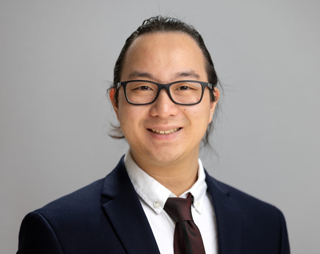 Anson smiles. Anson wears black glasses and a suit.