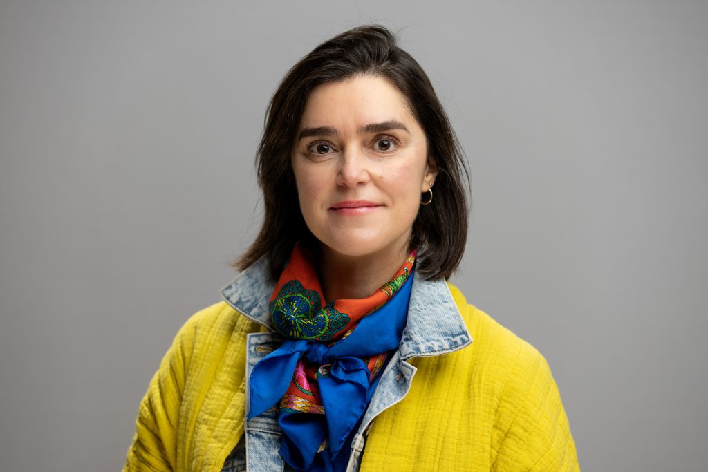 Amaya Atucha smiles for a professional photo. She has short, brown hair and wears yellow.