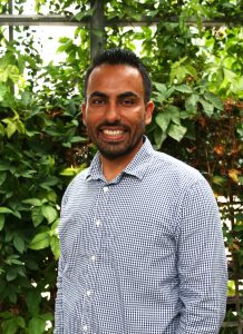 Raj smiles and wears a button up. A greenhouse is in the background.