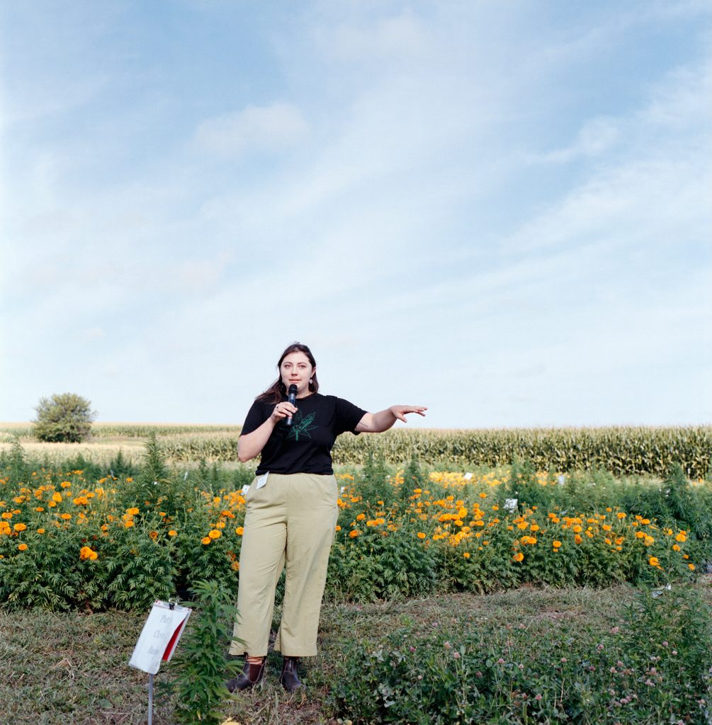 A person speaks into a microphone in a field of hemp and orange flowers.