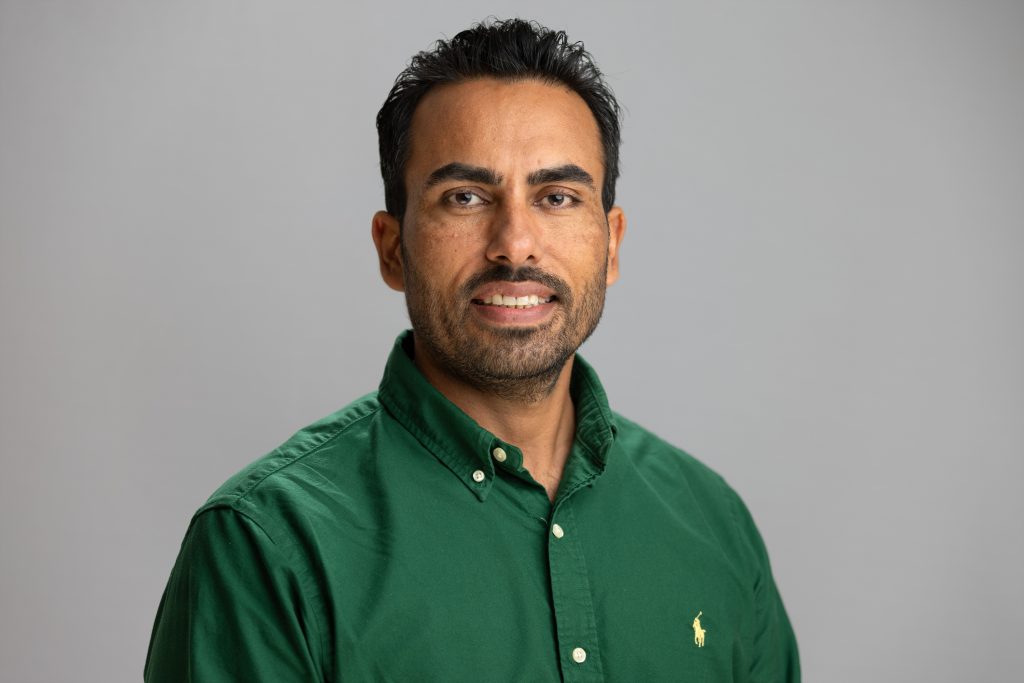 Raj smiles. He has short, black hair and wears a forest green button up.