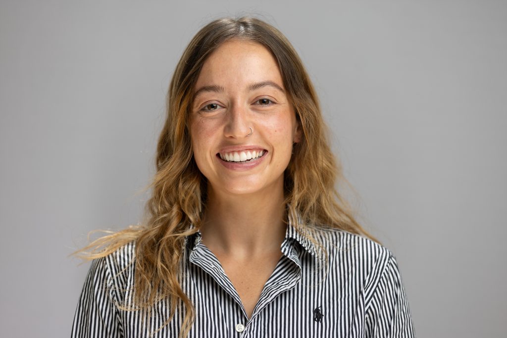 Sarah smiles. She has long, light brown hair and wears a striped button up.