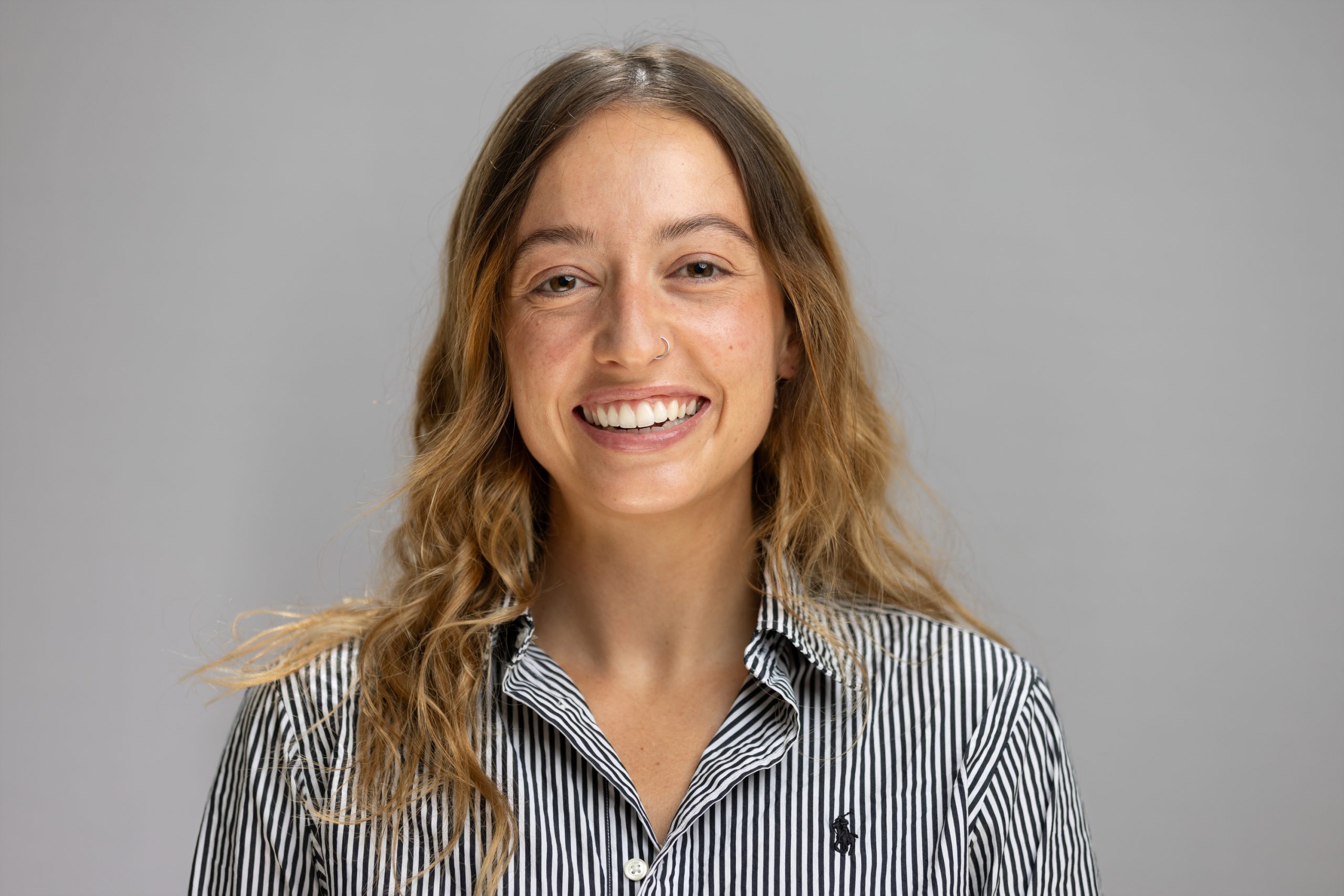 Sarah smiles. She has long, light brown hair and wears a striped button up.