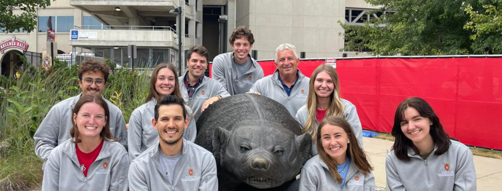 Ten people smile and wear matching gray shirts. They take a group photo around a badger statue.
