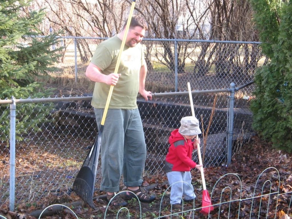 An adult and a toddler rake leaves together.