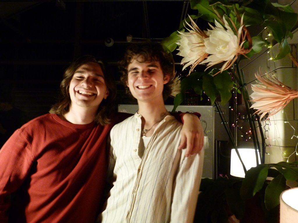 Two people smile next to large, beautiful flower blossoms.