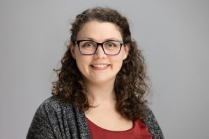 LauraLee smiles. She has brown, curly hair and wears a grey cardigan and red blouse.