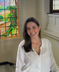 Livia smiles and wears a white blouse. In the background is a stained glass window.