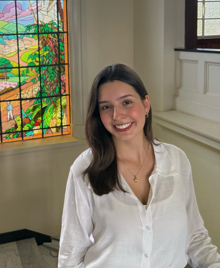 Livia smiles and wears a white blouse. In the background is a stained glass window.