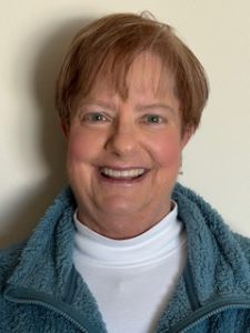 Amy smiles. She has brown hair and wears a white sweater and blue jacket.