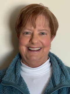 Amy smiles. She has brown hair and wears a white sweater and blue jacket.