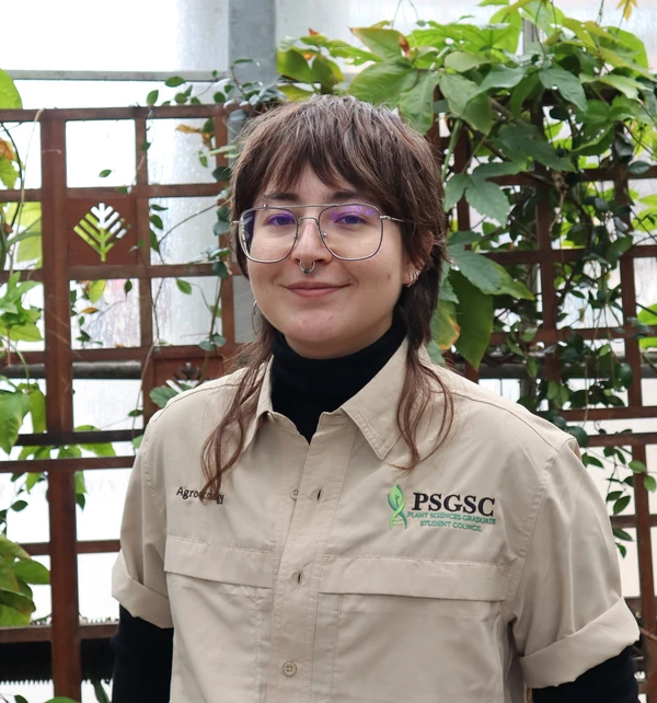 Andy smiles and has brown hair with curls. Andy wears a tan PSGSC polo and stands in a greenhouse.