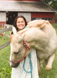 Sarah smiles and hugs a white horse in a pasture.
