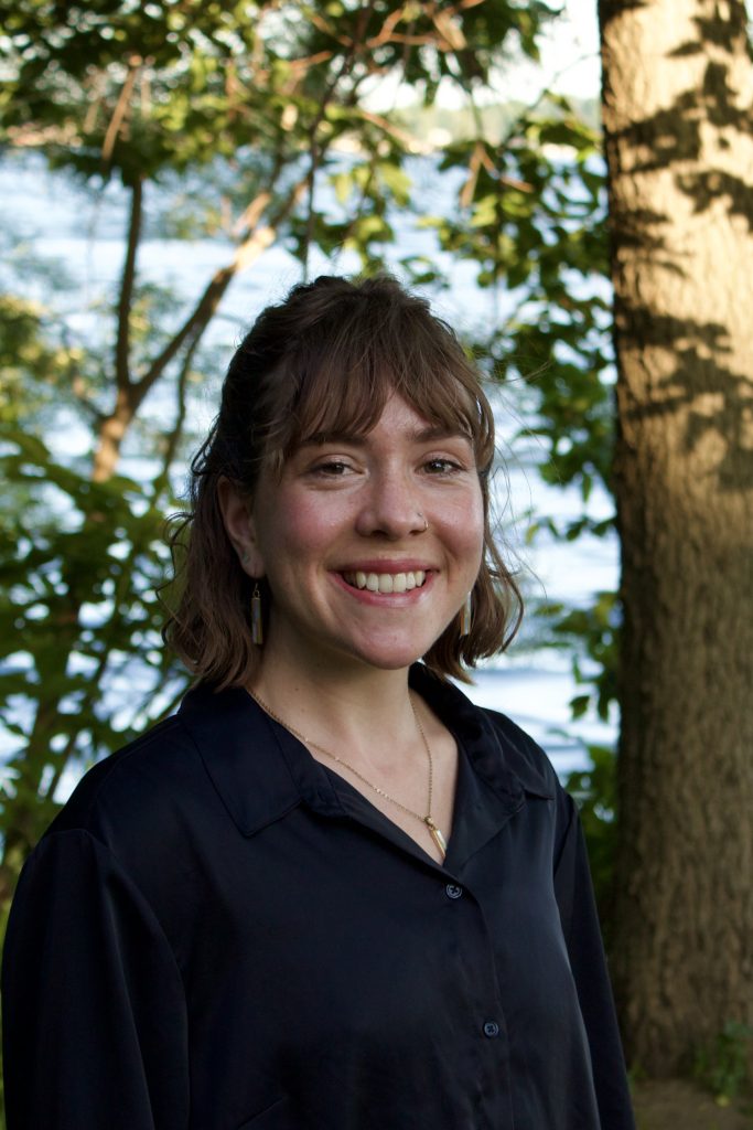 Jenny smiles. Jenny has short hair and stands outside in the woods.