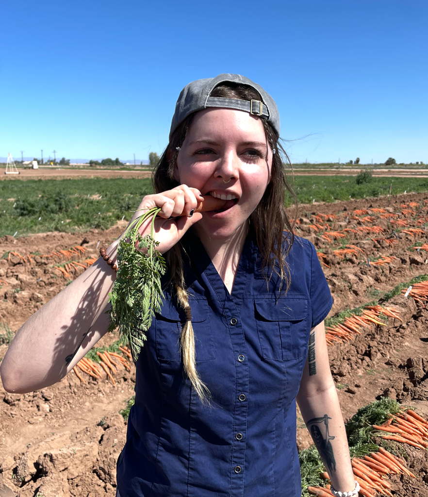 Avelina works in the field. Behind her, carrots lay in rows. She bites into a large carrot.