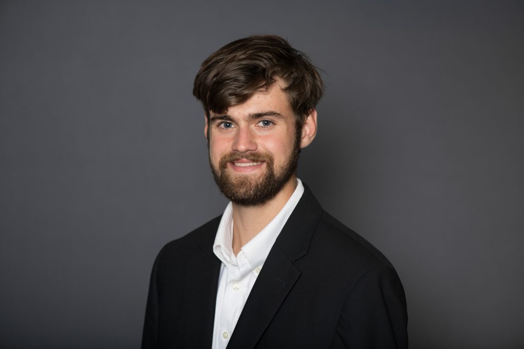 Joseph smiles for a professional portrait. Joseph has short, dark hair and wears a white button up.