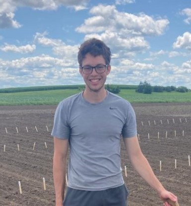 Hudson smiles on a sunny day in the field. Hudson has short hair and wears a grey blue shirt.