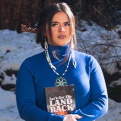 Tanikwah stands outside in the winter and holds a book with the title "Let's Get the Land Back."