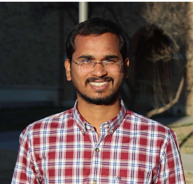Sai Ram Nagalla smiles for a photo. He wears a plaid shirt, glasses, and has short, dark hair.