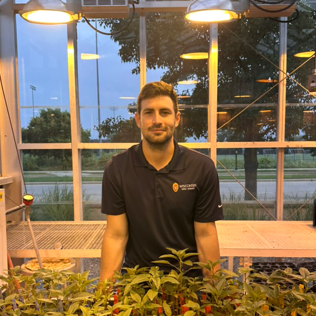 Guilherme smiles in a greenhouse. He wears a black polo with a UW-Madison logo on it.