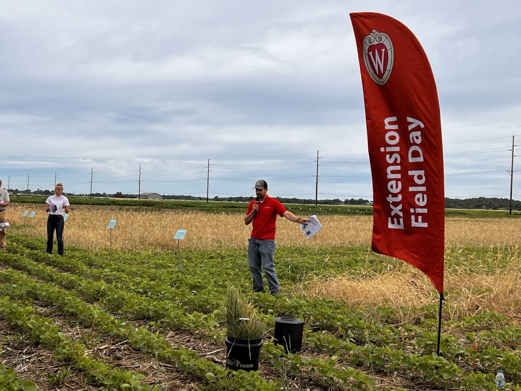 Guilherme stands in a field with a red sign that says "Extension Field Day." He wears a red polo and holds a microphone.