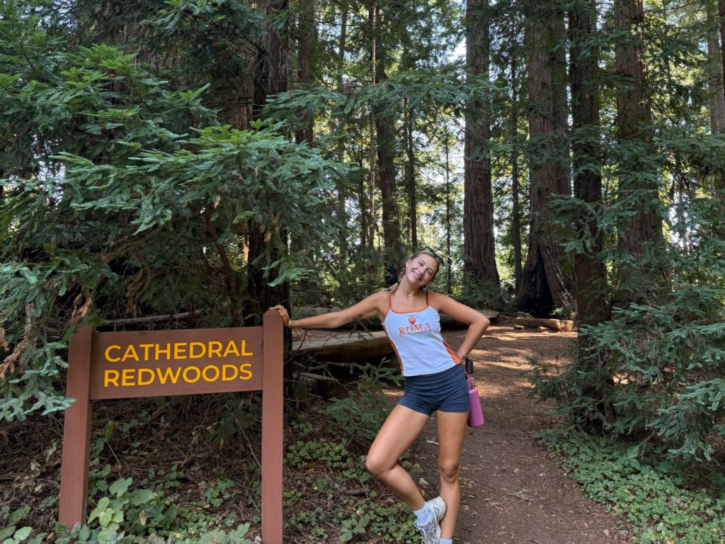 Hanf smiles and poses next to a sign that says "Cathedral Redwoods."