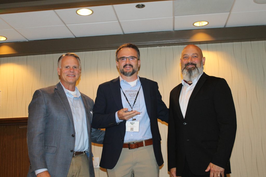 Three people in suits smile for a photo. The person in the middle holds an award.