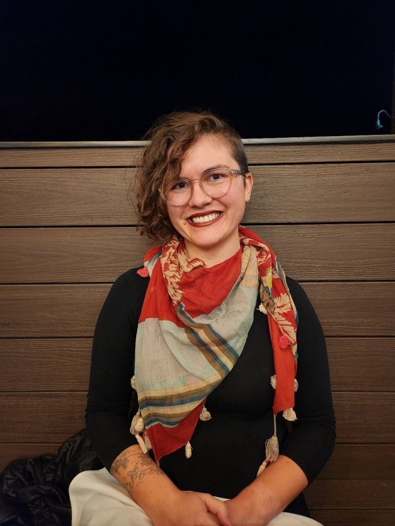 Mikki smiles. Mikki has short, curly hair and wears a scarf.